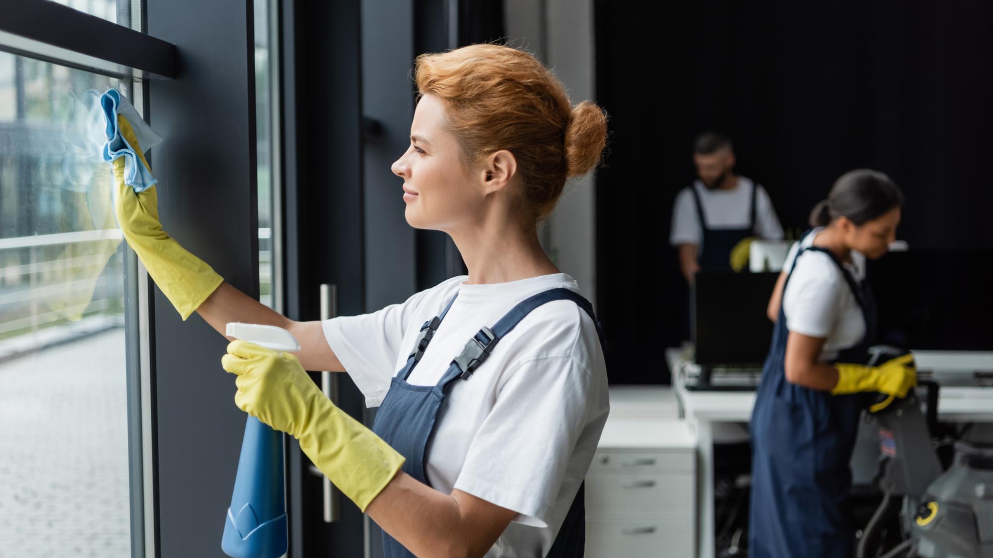A woman wearing a white shirt and blue overalls, with yellow cleaning gloves, is wiping a window with a cloth. In the background, two individuals are seen: one is vacuuming and the other is working at a desk. The setting appears to be an office space.